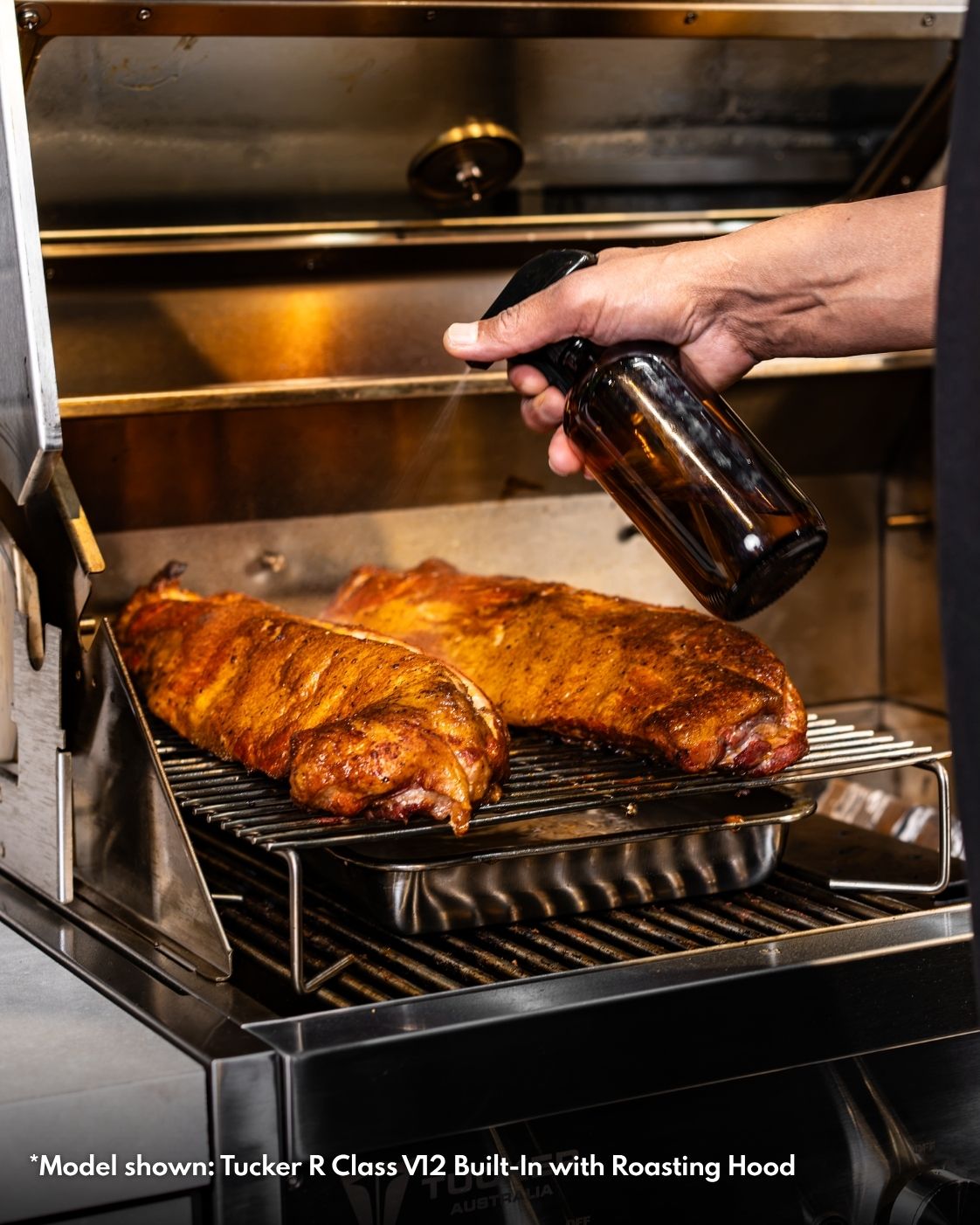 Texas-style pork ribs being spritzed on a roasting rack above a drip pan inside a Tucker R Class BBQ