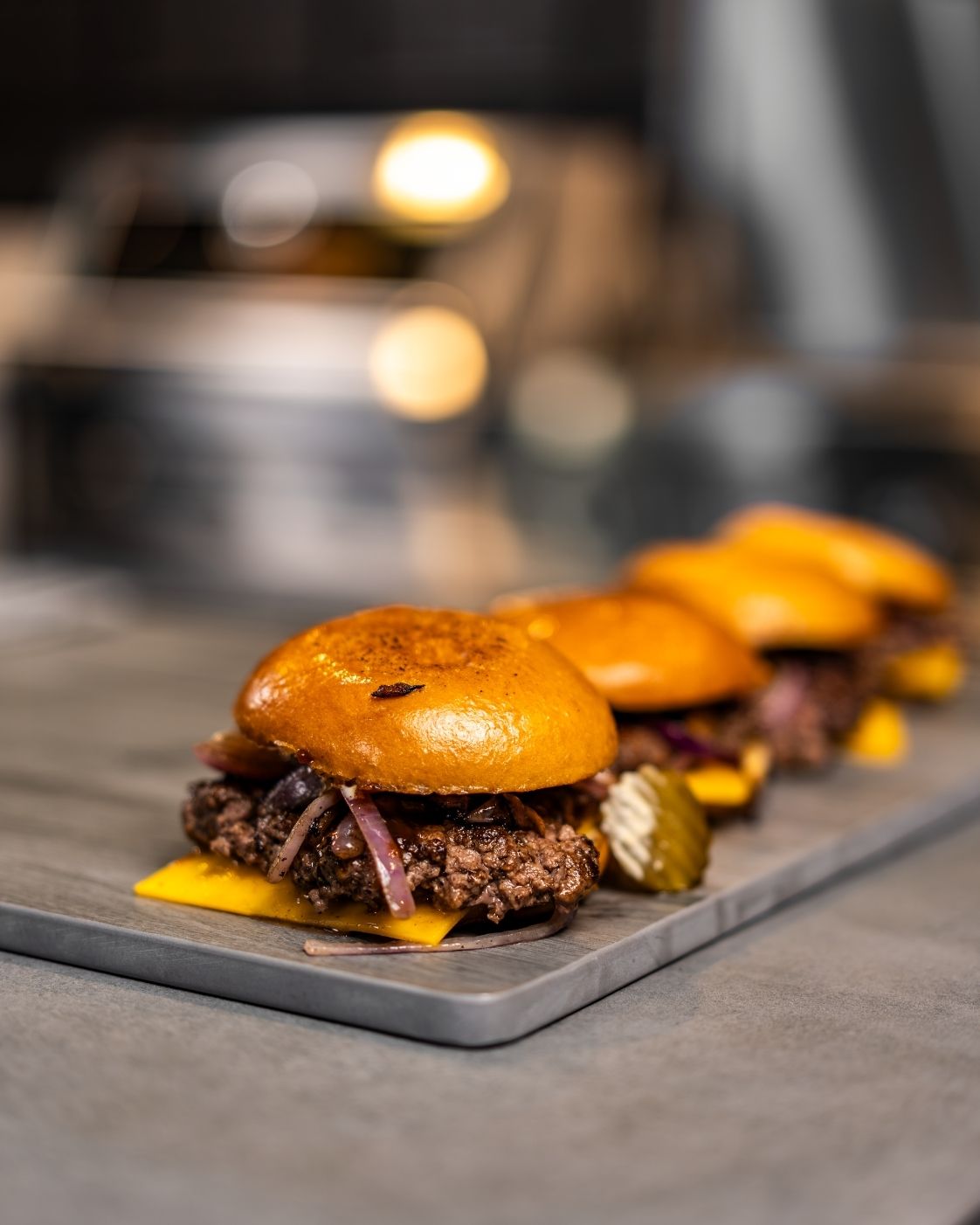 Beef cheeseburger sliders with onion and pickles on a serving board, with a Tucker R Class BBQ behind