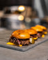 Beef cheeseburger sliders with onion and pickles on a serving board, with a Tucker R Class BBQ behind